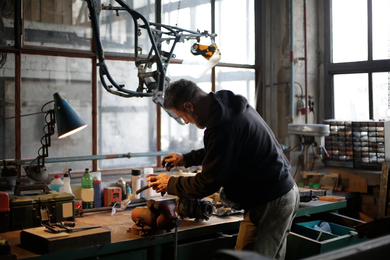 services-02 Back view of unrecognizable male mechanic in workwear standing near workbench while fixing broken metal detail in lamplight in modern workshop